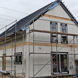 DEWERIS HOLDING PROSTA SPÓŁKA AKCYJNA - Budowa domu szkieletowego: widok na elewację z oknami i rusztowaniami. Widoczny fragment dachu z membraną. Etap prac przed ociepleniem.