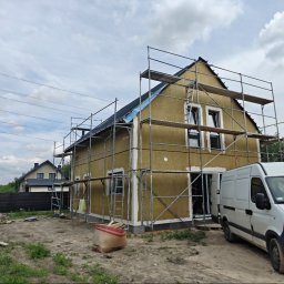 DEWERIS HOLDING PROSTA SPÓŁKA AKCYJNA - Dom jednorodzinny w trakcie ocieplania, otoczony rusztowaniem. Widoczny biały samochód dostawczy i materiały budowlane na posesji. W tle drugi dom.