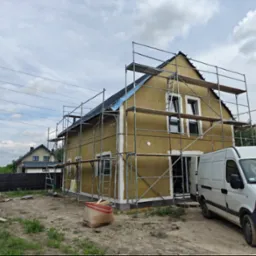 Dom jednorodzinny w trakcie ocieplania, otoczony rusztowaniem. Widoczny biały samochód dostawczy i materiały budowlane na posesji. W tle drugi dom.