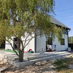 Nowoczesny, biały dom z szarą elewacją, widoczny z boku, z dużym drzewem na pierwszym planie i tarasem na podeście. Widoczne narzędzia i pranie sugerują prace wykończeniowe.