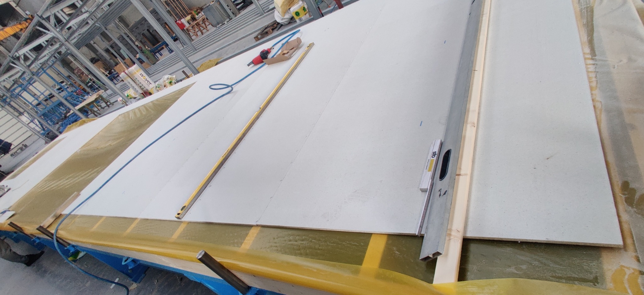 Przygotowywanie paneli ściennych w hali produkcyjnej, widoczne poziomice i narzędzia pneumatyczne, konstrukcja stalowa w tle.