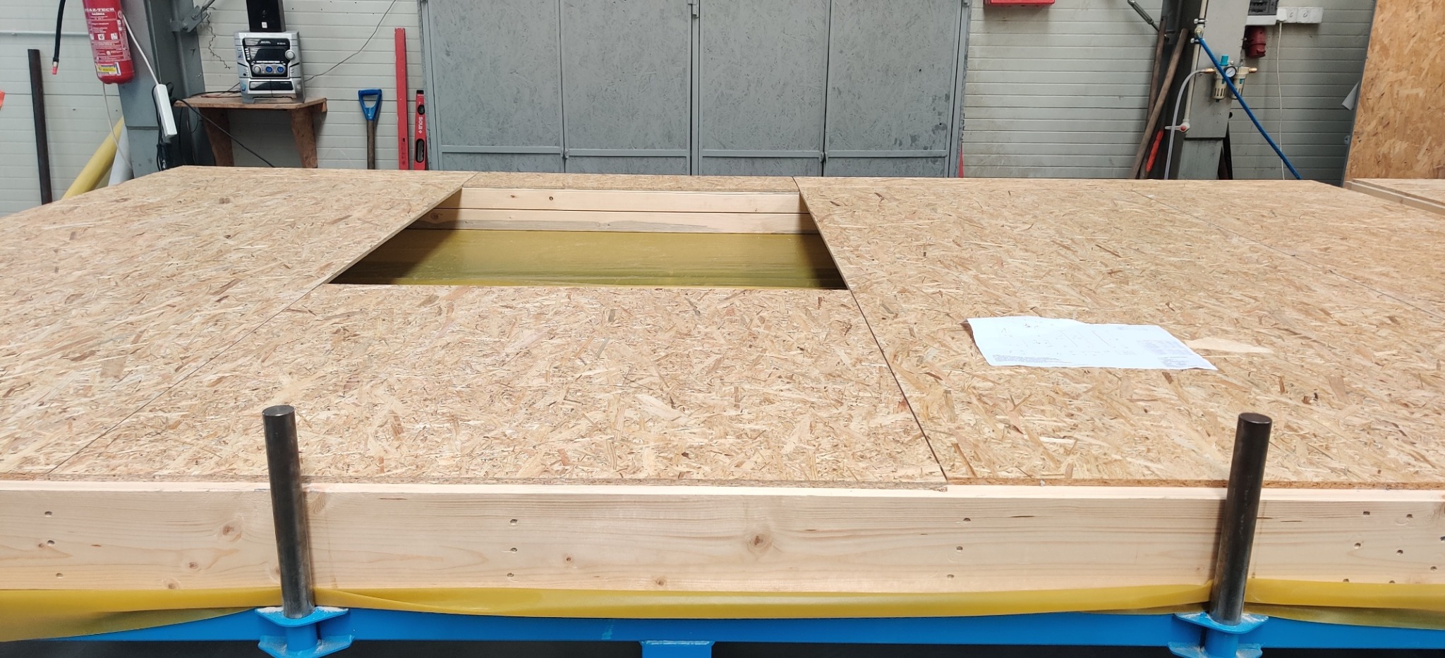 Fragment konstrukcji podłogi z płyty OSB z otworem, umieszczonej na niebieskiej ramie, zabezpieczonej żółtą folią, z widocznym planem na papierze.