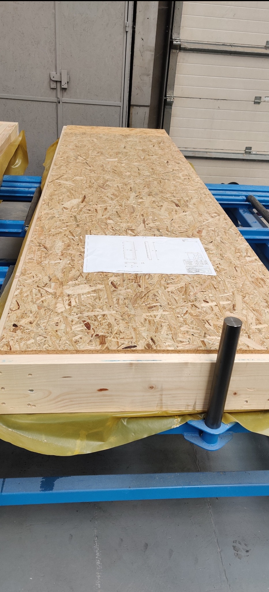 Panel OSB z drewnianą ramą, zabezpieczony folią, leżący na niebieskiej konstrukcji, z widocznym planem technicznym na górnej powierzchni.