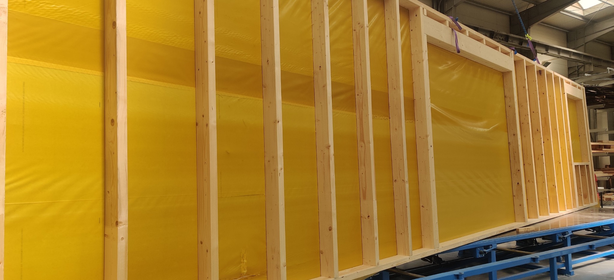 Fragment ściany szkieletowej z żółtą membraną wewnątrz ramy, widoczna konstrukcja drewniana, przygotowana do transportu na niebieskim wózku transportowym w hali produkcyjnej.