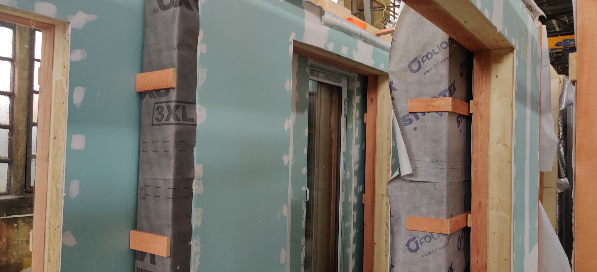 Wnętrze domu w trakcie budowy: ściany z płyt gipsowo-kartonowych, częściowo pokryte folią izolacyjną z nadrukiem i drewnianymi listwami mocującymi, widoczne ramy okienne i elementy konstrukcyjne.