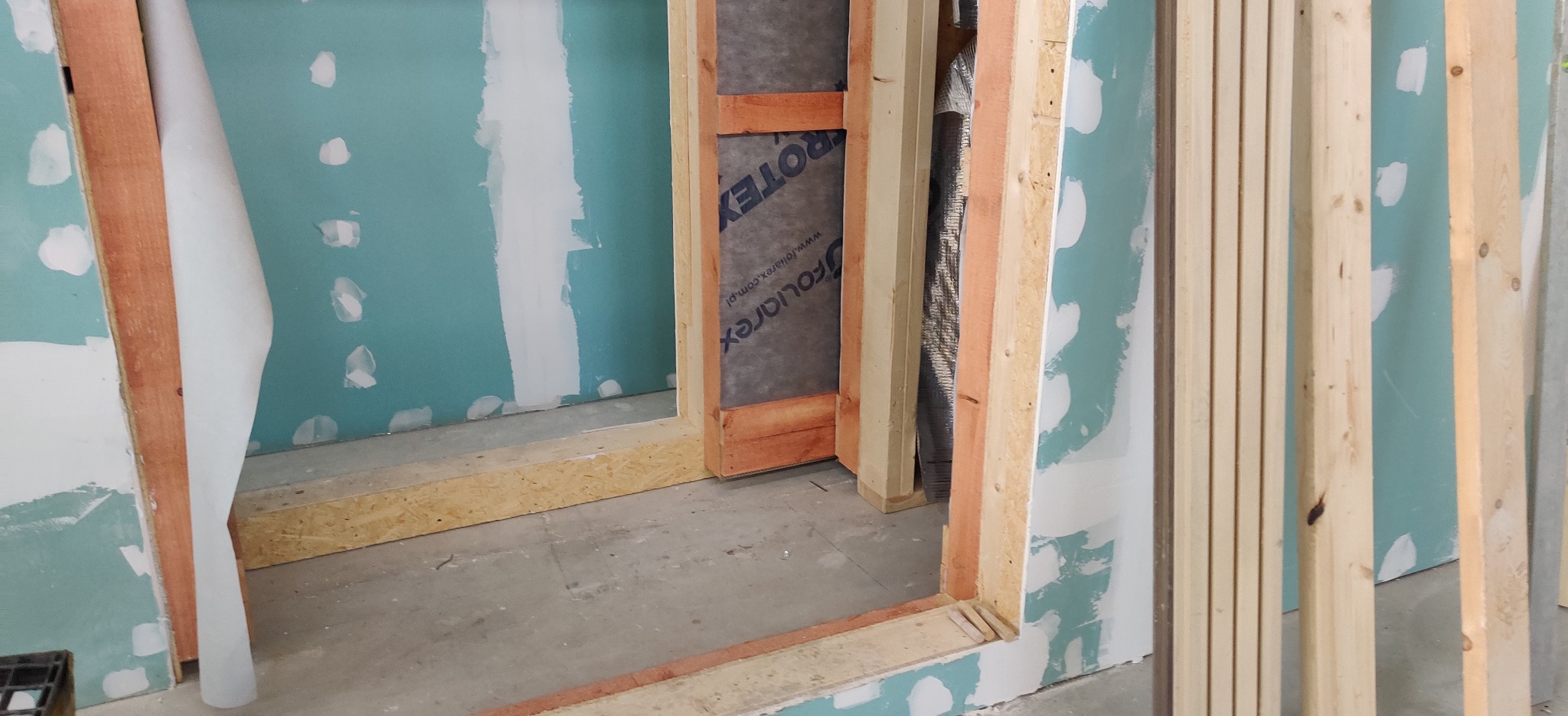 Wnętrze w trakcie budowy, widoczne drewniane ramy drzwi, fragment ściany z płyt gipsowo-kartonowych z widocznymi łączeniami, materiał izolacyjny z folią aluminiową oraz stos drewnianych listew