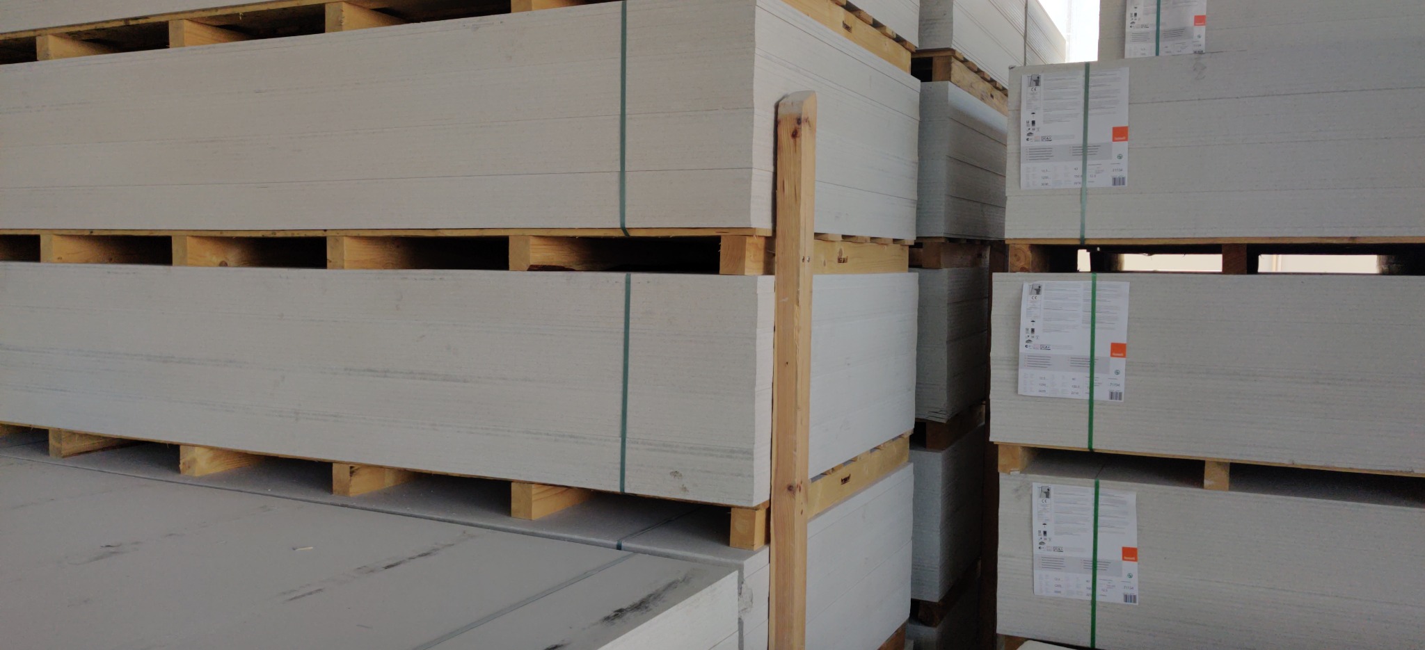 Składowane płyty gipsowo-kartonowe na paletach, zabezpieczone zielonymi taśmami, z widocznymi etykietami producenta.