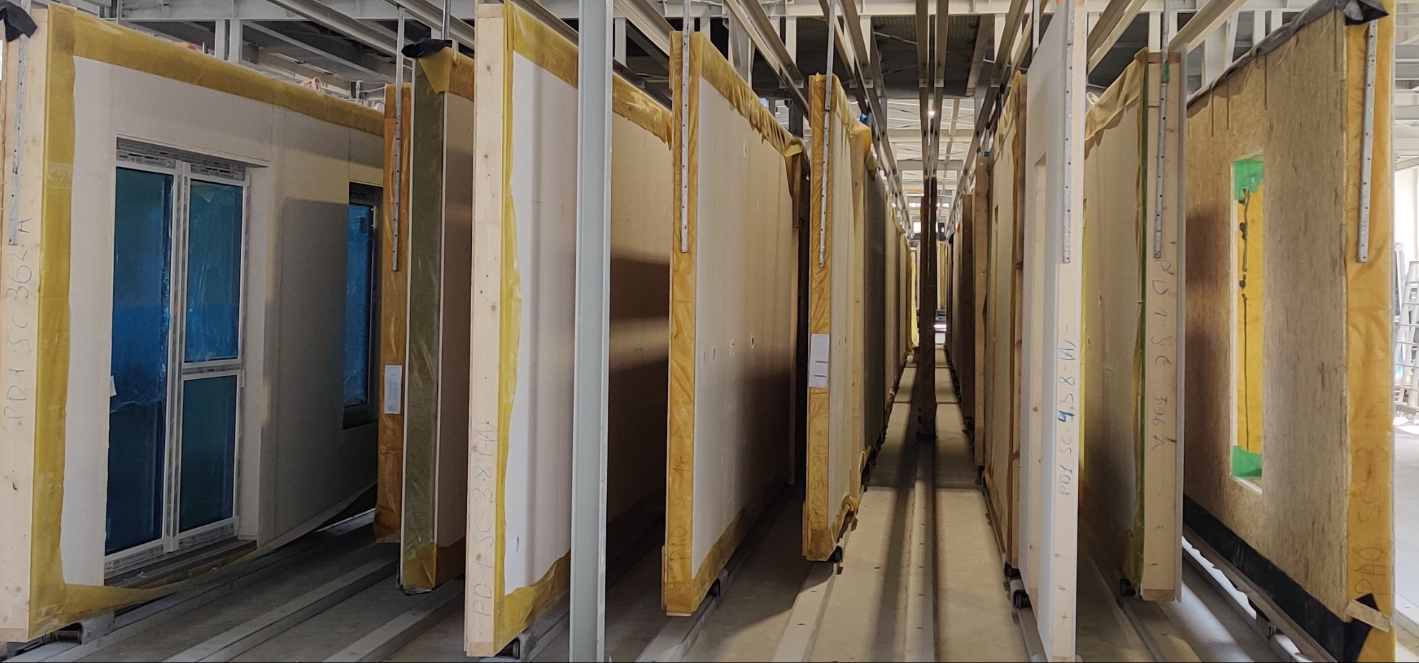 Magazyn prefabrykowanych ścian z oknami, widoczne izolacje i oznaczenia kodowe, przygotowane do montażu.