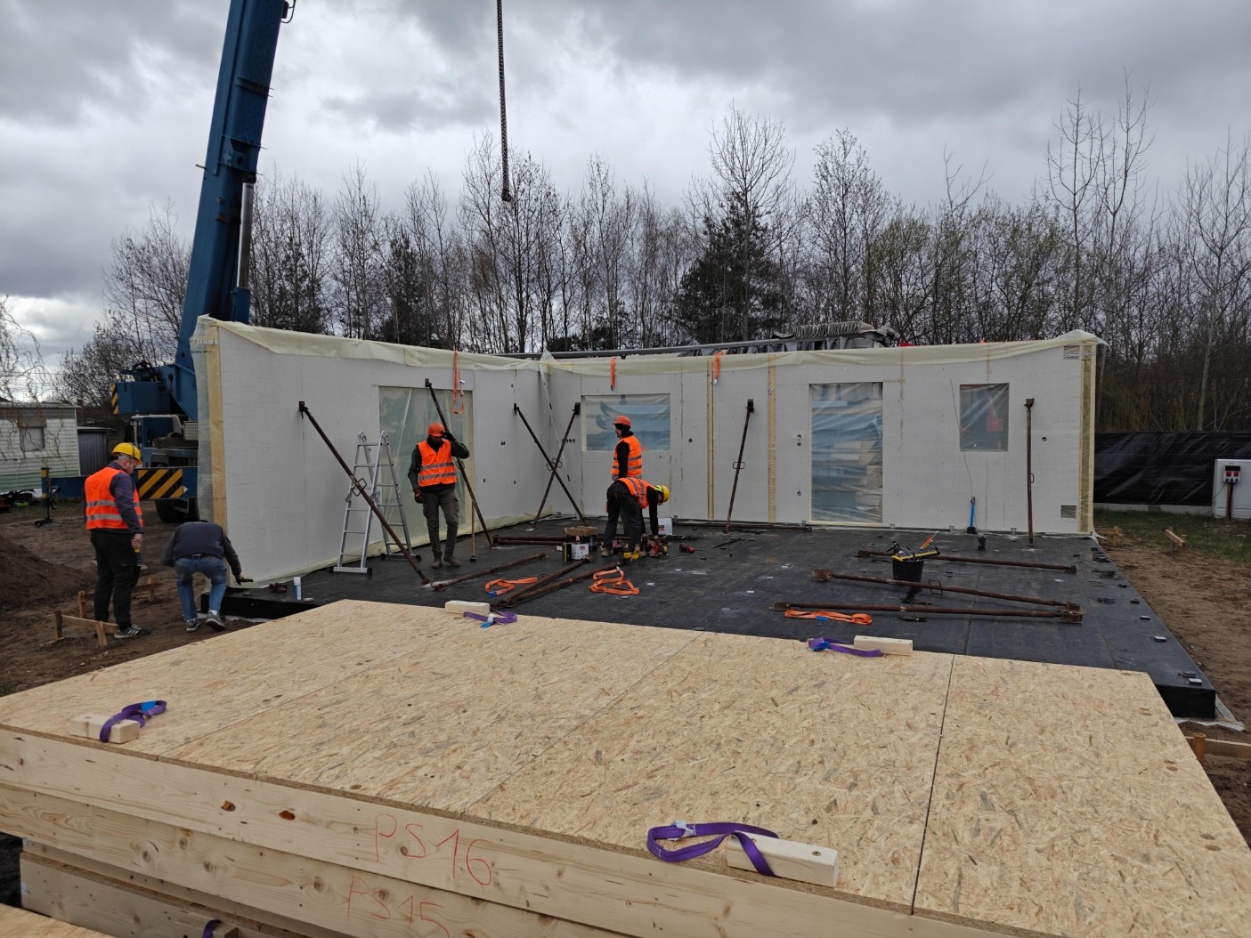 Montaż ścian domu modułowego z użyciem dźwigu. Widoczni pracownicy w kamizelkach, elementy konstrukcyjne i narzędzia. Etap budowy z widoczną konstrukcją podłogi.