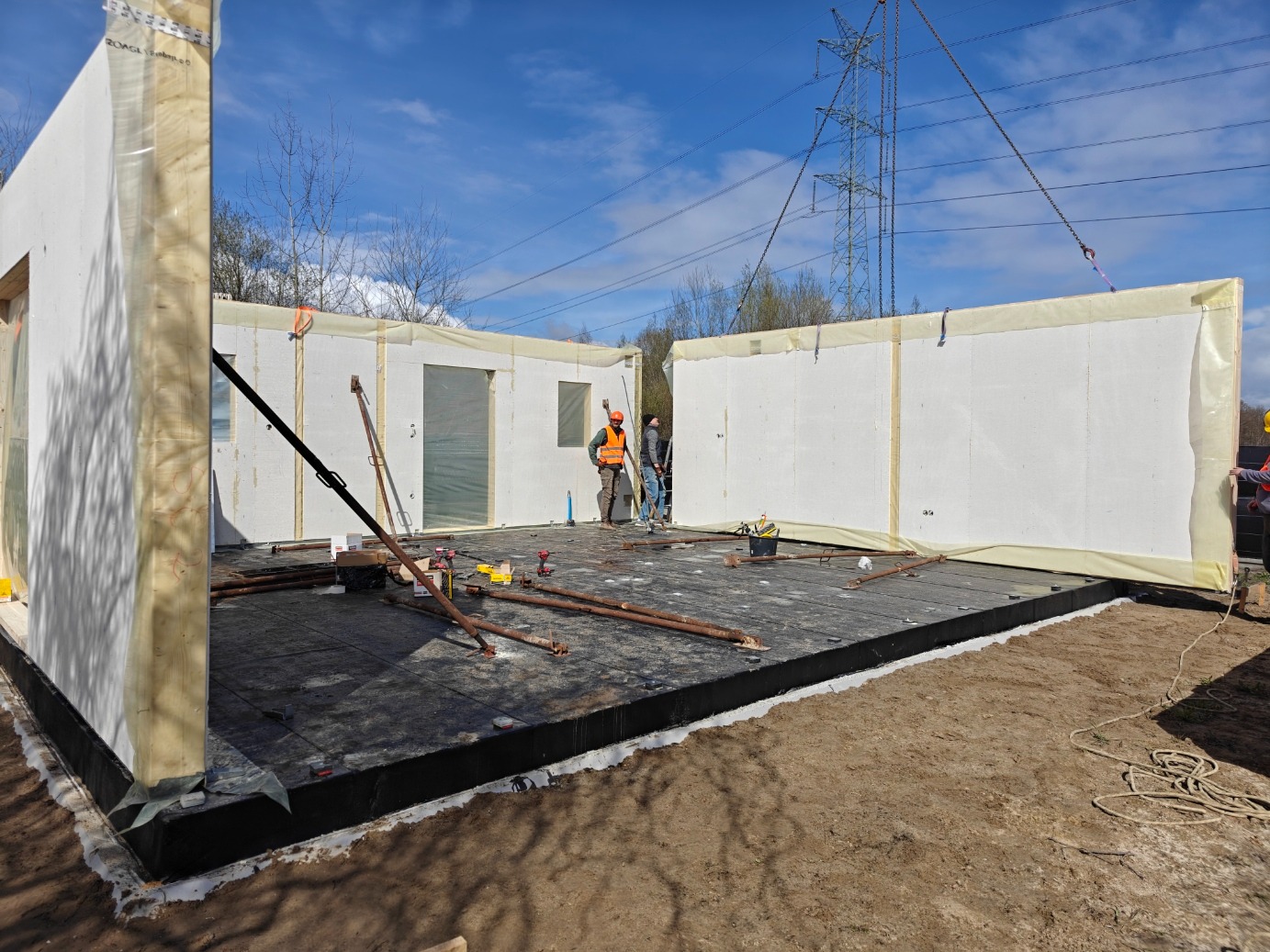 Montaż modułowej konstrukcji domu: robotnicy w kamizelkach montują ściany na przygotowanym fundamencie, widoczne narzędzia i elementy konstrukcyjne.