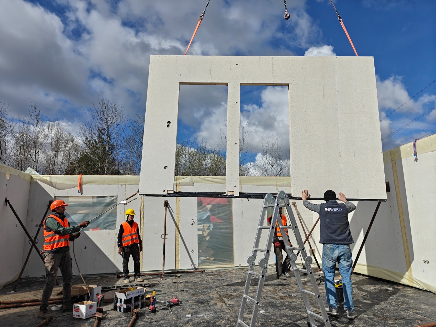 Montaż segmentu domu modułowego z oknami przez ekipę budowlaną. Element podnoszony dźwigiem, pracownicy w kaskach i kamizelkach. Widoczne narzędzia i drabina.