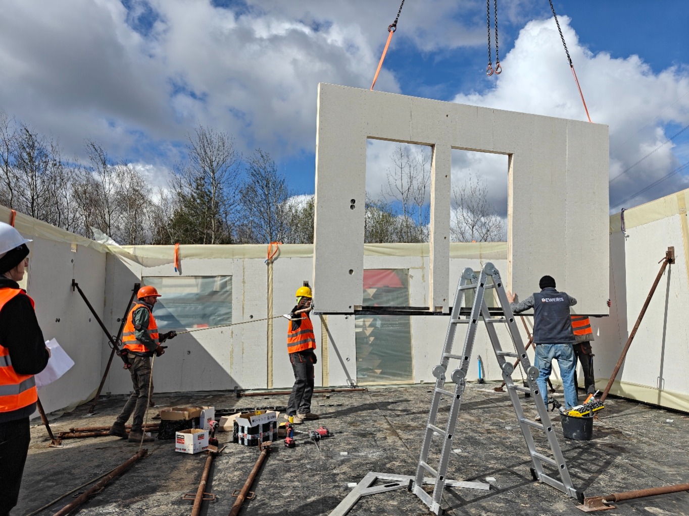Montaż ściany z dwoma oknami na budowie domu modułowego. Pracownicy w kaskach i kamizelkach, drabina, narzędzia. Jasne, pochmurne niebo w tle.
