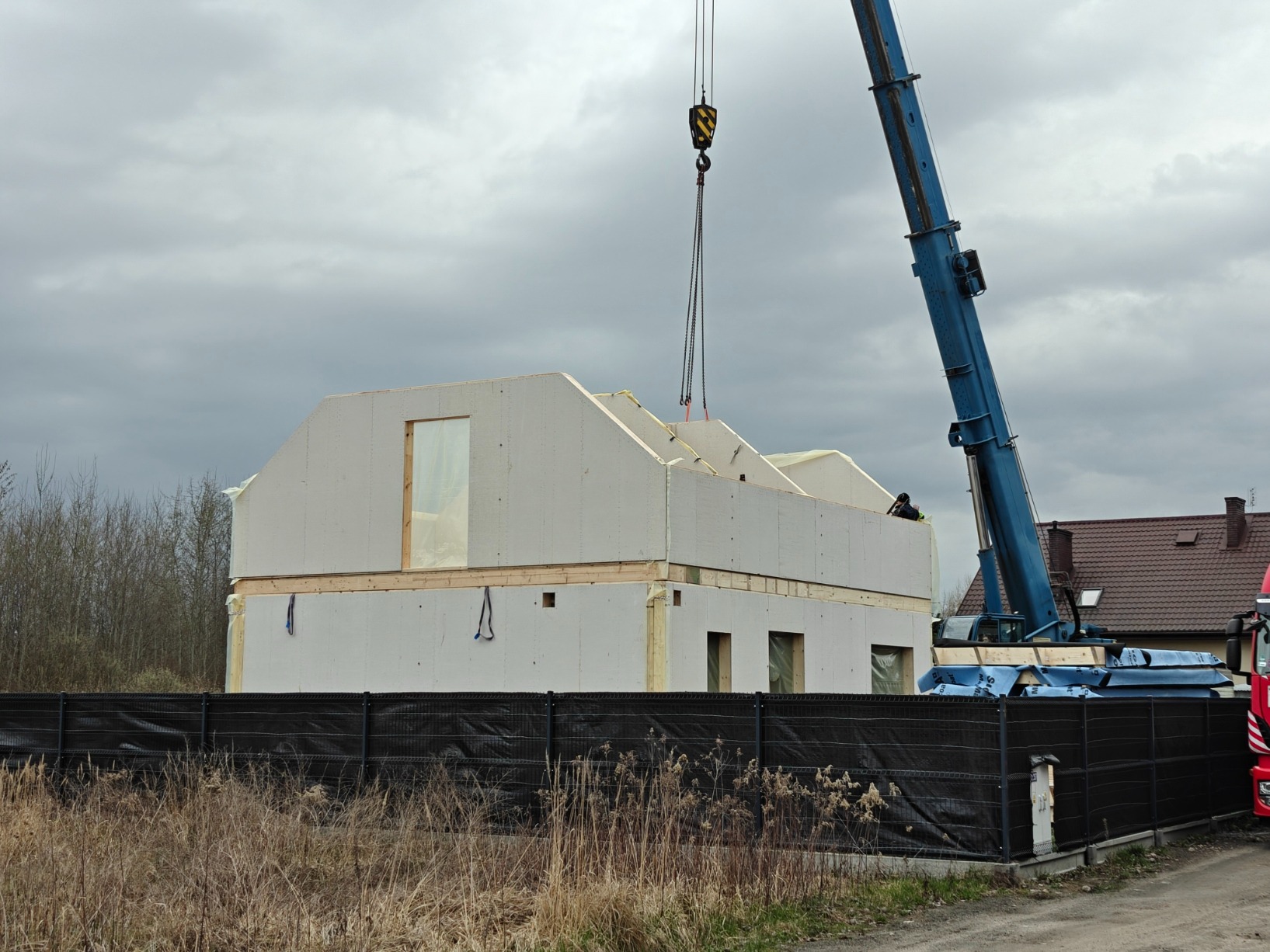 Montaż segmentu domu szkieletowego za pomocą dźwigu na tle pochmurnego nieba. Widoczne elementy konstrukcyjne i zabezpieczenia.