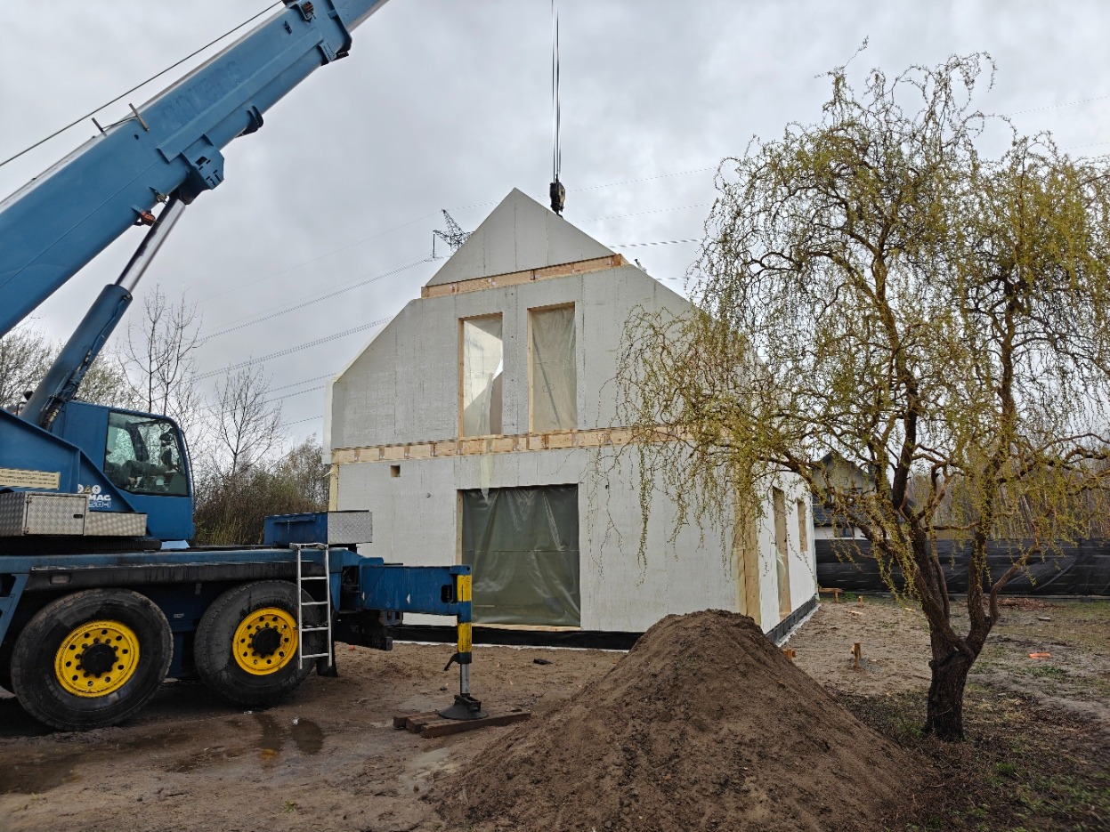 Montaż domu modułowego z użyciem dźwigu. Widoczne panele ścian i konstrukcja dachu w surowym stanie. Na pierwszym planie dźwig i hałda ziemi.