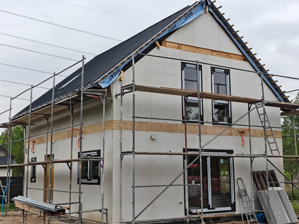 Budowa domu szkieletowego: widok na elewację z oknami i rusztowaniami. Widoczny fragment dachu z membraną. Etap prac przed ociepleniem.