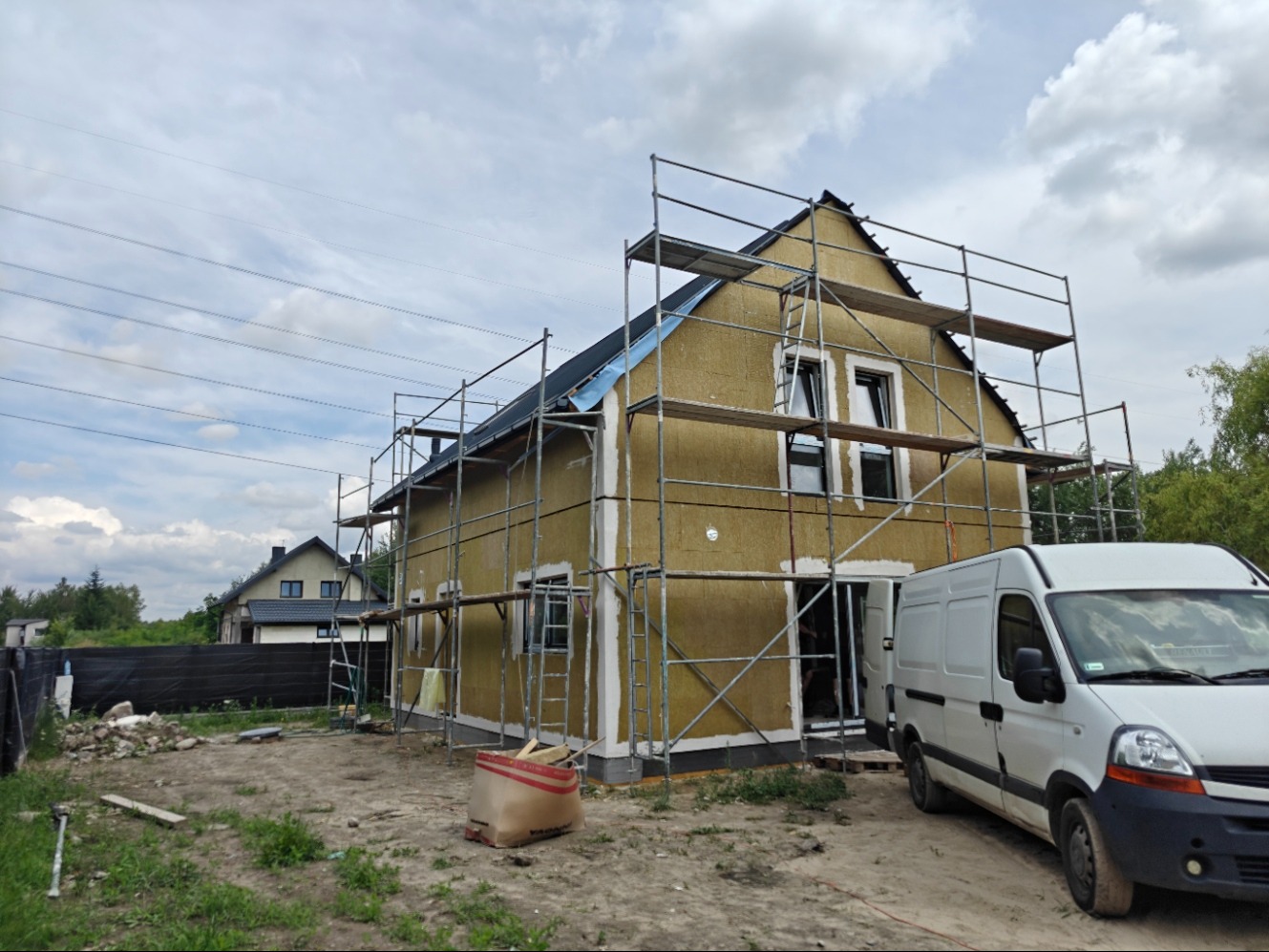 Dom jednorodzinny w trakcie ocieplania, otoczony rusztowaniem. Widoczny biały samochód dostawczy i materiały budowlane na posesji. W tle drugi dom.