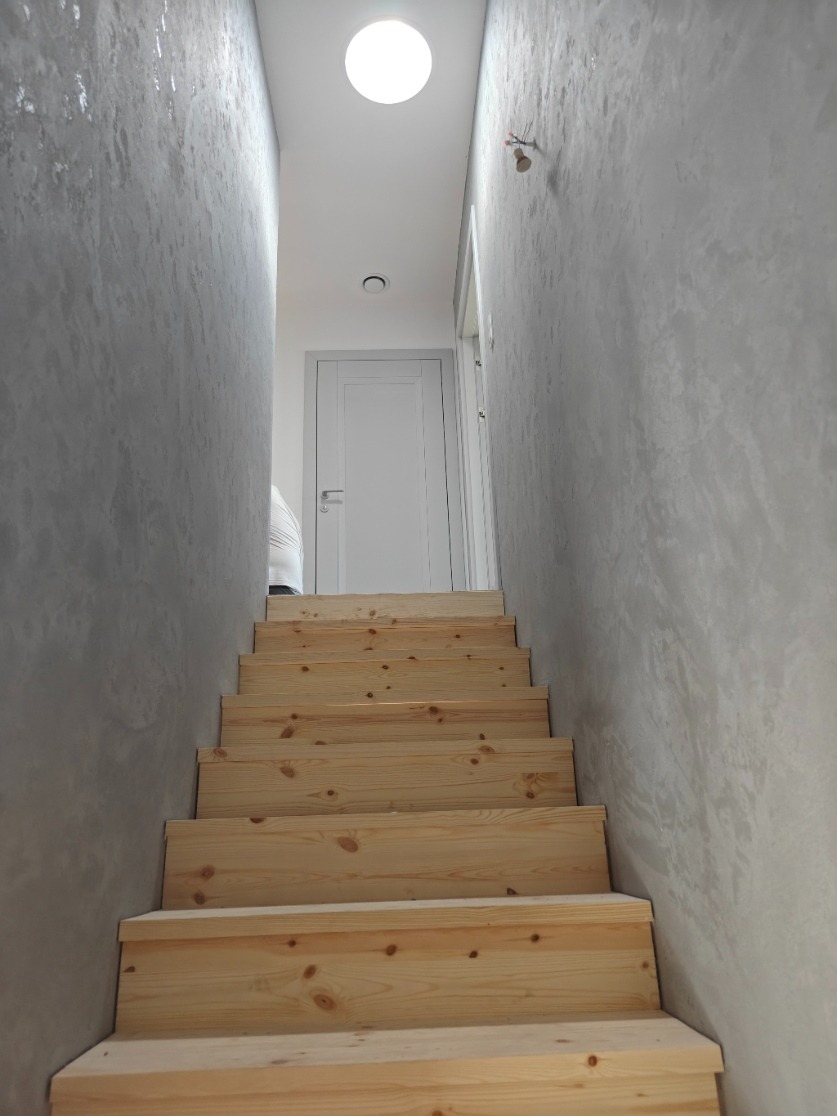 Klatka schodowa z surowego drewna prowadzi do białych drzwi. Ściany szare, fakturowane. Okrągłe światło sufitowe. Minimalistyczny, nowoczesny design.