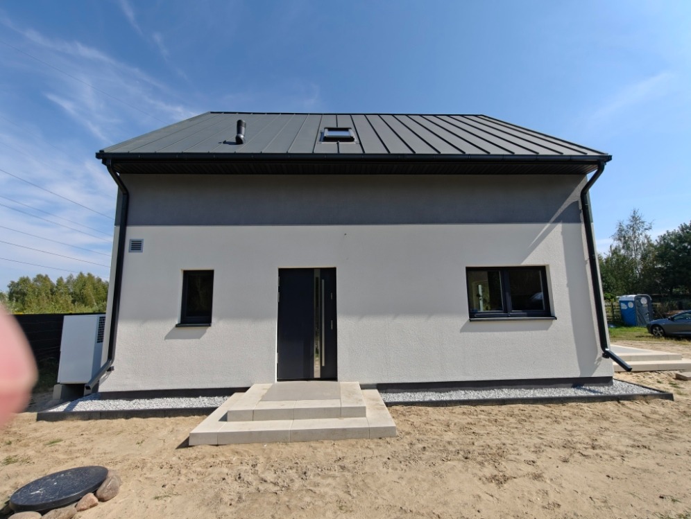Nowoczesny, parterowy dom z szarą elewacją i dachem z blachy na tle błękitnego nieba. Widoczne schody wejściowe i zagospodarowany teren wokół budynku.