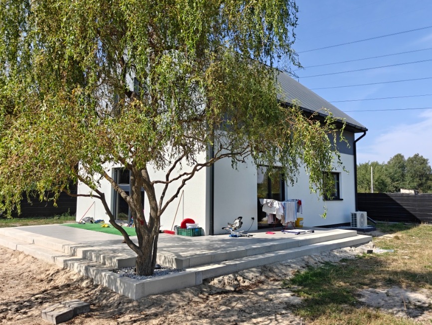Nowoczesny, biały dom z szarą elewacją, widoczny z boku, z dużym drzewem na pierwszym planie i tarasem na podeście. Widoczne narzędzia i pranie sugerują prace wykończeniowe.