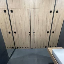 Serhii Hubrii SH ART - Szafa wnękowa z drewnopodobnymi frontami i czarnymi detalami, wkomponowana w przestrzeń łazienki z szarą podłogą i umywalką na drewnianym blacie.