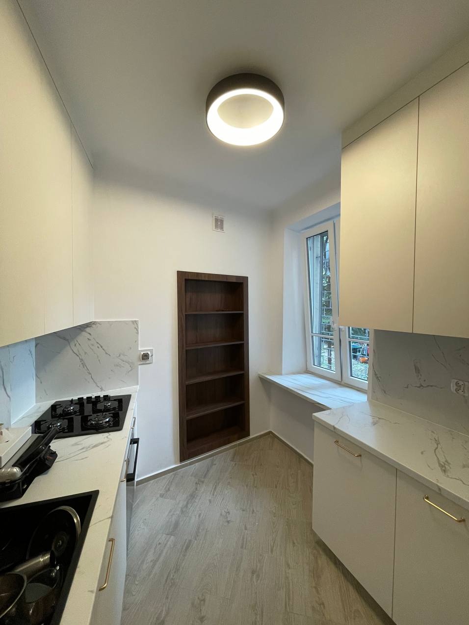 Nowoczesna kuchnia z białymi szafkami, marmurowym blatem, wbudowaną ciemnobrązową półką na książki i okrągłą lampą sufitową.