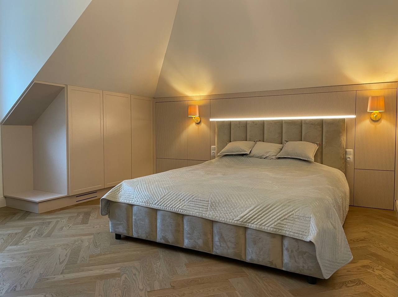 Sypialnia na poddaszu z zabudową meblową w jasnych odcieniach, łóżko z tapicerowanym wezgłowiem i oświetleniem LED, parkiet ułożony w jodełkę.