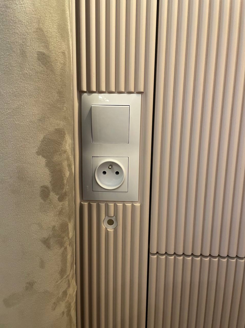 Fragment beżowej ściany z pionowymi żłobieniami, wbudowany włącznik światła i gniazdko elektryczne, po lewej stronie fragment beżowej ściany.