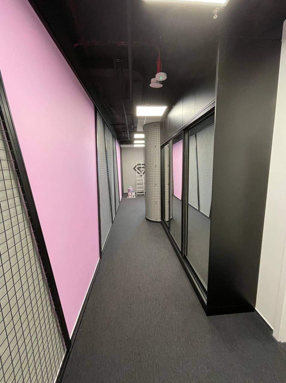 Długi korytarz z szafami przesuwnymi o czarnych ramach, różowymi ścianami i czarną podłogą, widoczna drabina i puszki z farbą w oddali.