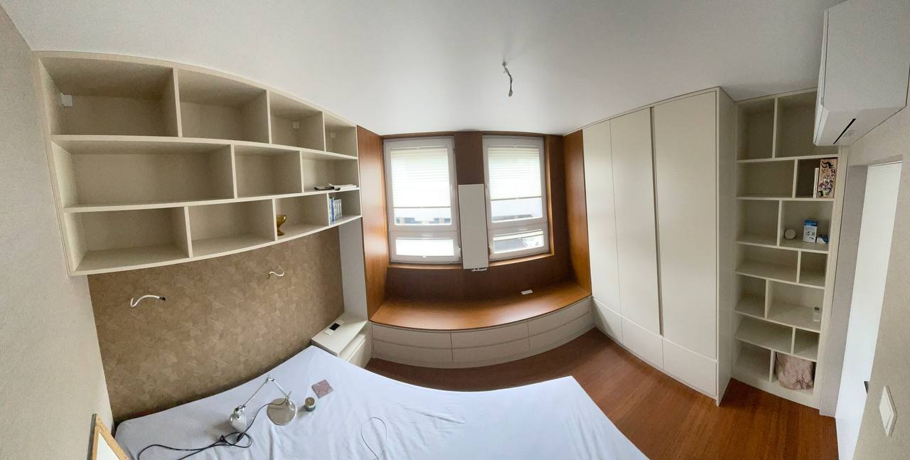 Pokój z łóżkiem, zabudową szaf i półek w jasnych odcieniach, z siedziskiem przy oknie i drewnianą podłogą.