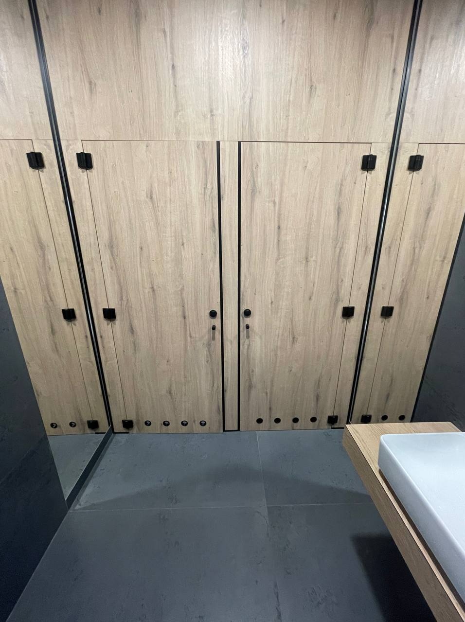 Szafa wnękowa z drewnopodobnymi frontami i czarnymi detalami, wkomponowana w przestrzeń łazienki z szarą podłogą i umywalką na drewnianym blacie.