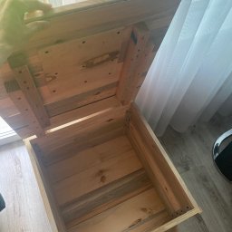 Krzysztof Czerwiński - Otwarta, surowa, drewniana skrzynia z widocznymi elementami mocującymi, stojąca przy oknie z białymi zasłonami.