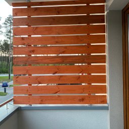 Krzysztof Czerwiński - Drewniana przesłona balkonowa z poziomych desek w ciepłym odcieniu brązu, zamocowana do ściany i balustrady balkonu za pomocą metalowych uchwytów, z widocznymi przerwami między deskami...
