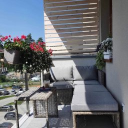 Krzysztof Czerwiński - Zacieniony balkon z rattanowym narożnikiem, stolikiem i drewnianą osłoną, kwiaty w donicach, widok na ulicę z samochodami.