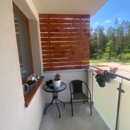 Krzysztof Czerwiński - Zadaszony balkon z drewnianą osłoną, szklaną balustradą, stolikiem kawowym, krzesłem z rattanu i kwiatami w doniczkach, widok na las w tle.