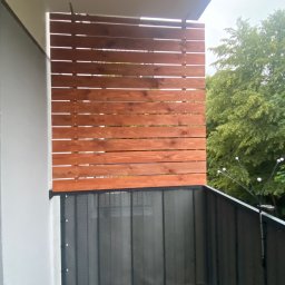 Krzysztof Czerwiński - Drewniana osłona balkonowa z poziomych desek w ciepłym odcieniu brązu, zamontowana nad metalową balustradą z siatki, widok z balkonu na zieleń drzew.