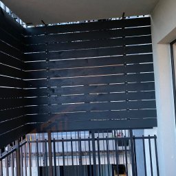 Krzysztof Czerwiński - Częściowy widok balkonu z poziomymi, ciemnymi deskami tworzącymi przesłonę, zamontowanymi na metalowej konstrukcji balustrady.