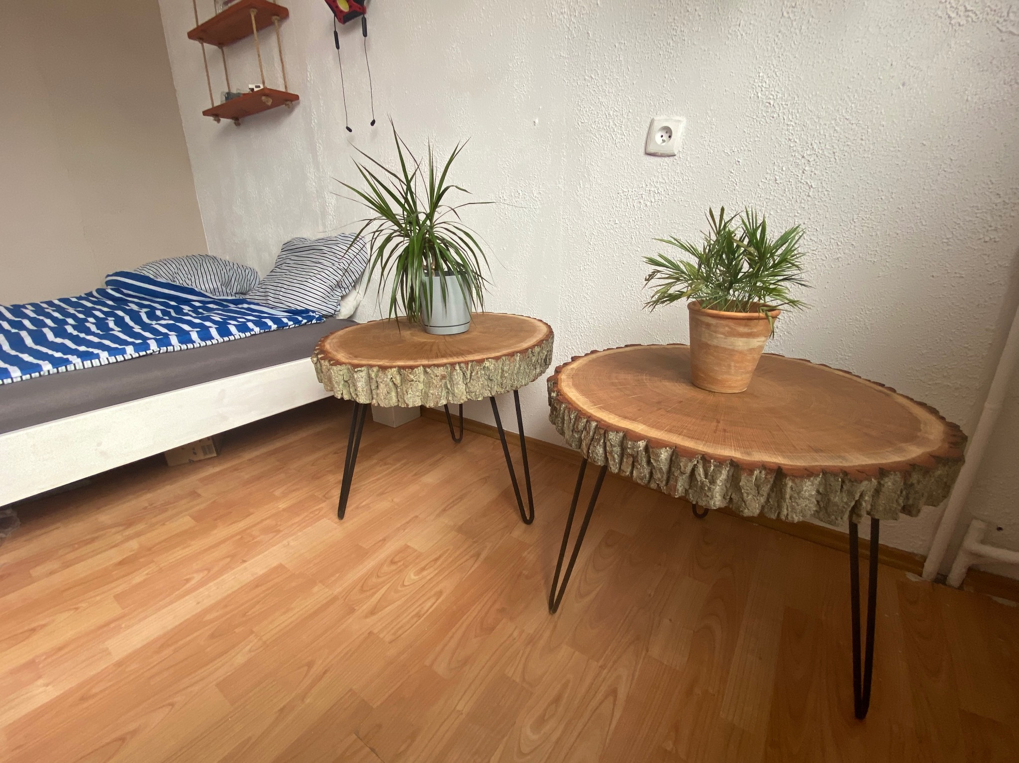 Dwa stoliki kawowe wykonane z plastrów drewna z korą i metalowymi nogami w kształcie litery V, na każdym z nich stoi doniczka z rośliną, w tle widoczne łóżko i półka.