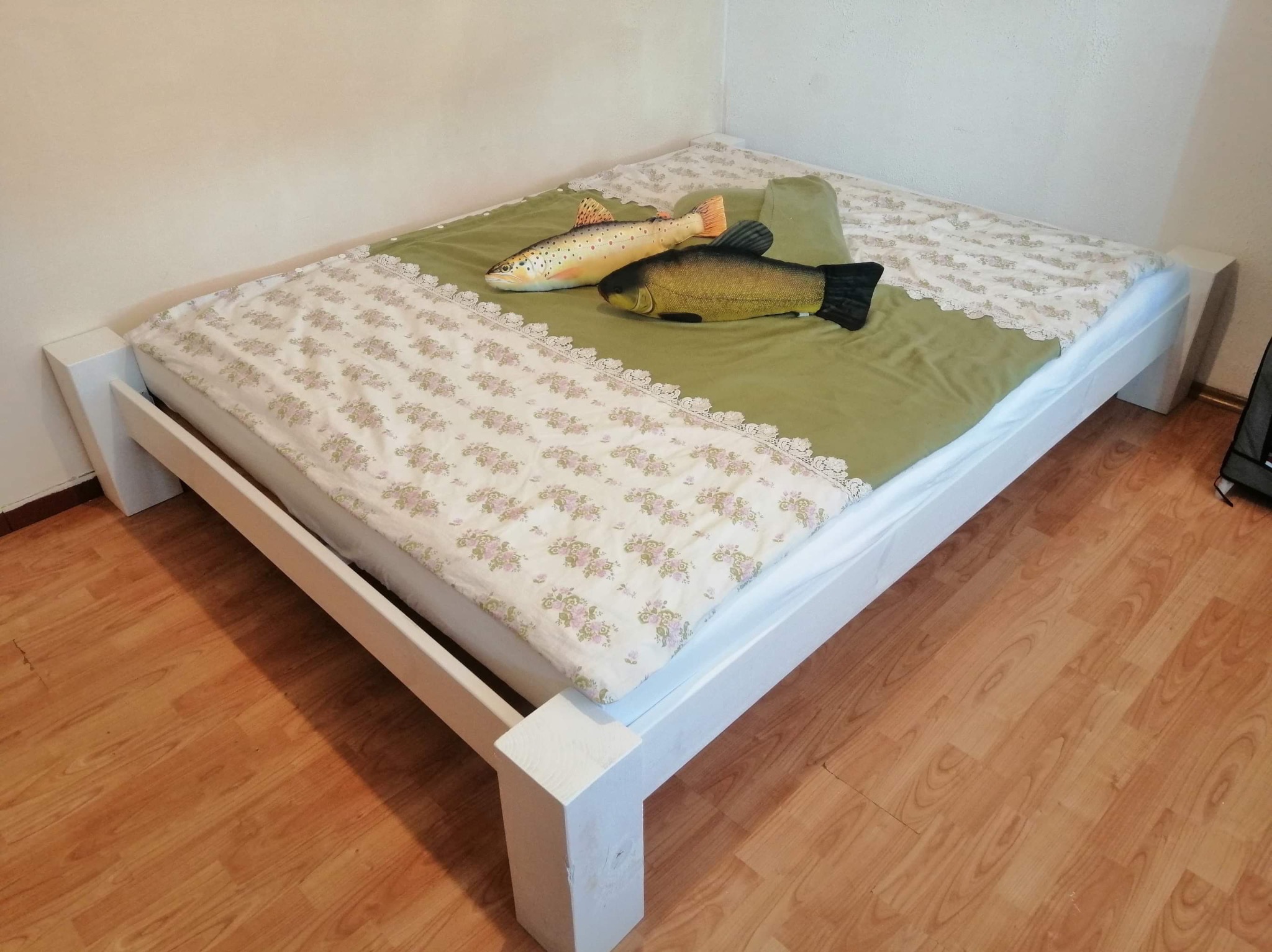 Proste, białe łóżko z drewnianą ramą na drewnianej podłodze, nakryte zieloną narzutą z koronkowym wykończeniem i wzorzystą pościelą, na której leżą dwie pluszowe ryby: pstrąg i lin.
