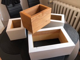 Zestaw czterech drewnianych skrzynek o różnych rozmiarach i wykończeniach, ustawionych na okrągłym stole z czarnym, wzorzystym blatem. Jedna skrzynka jest surowa, z widoczną strukturą drewna,...