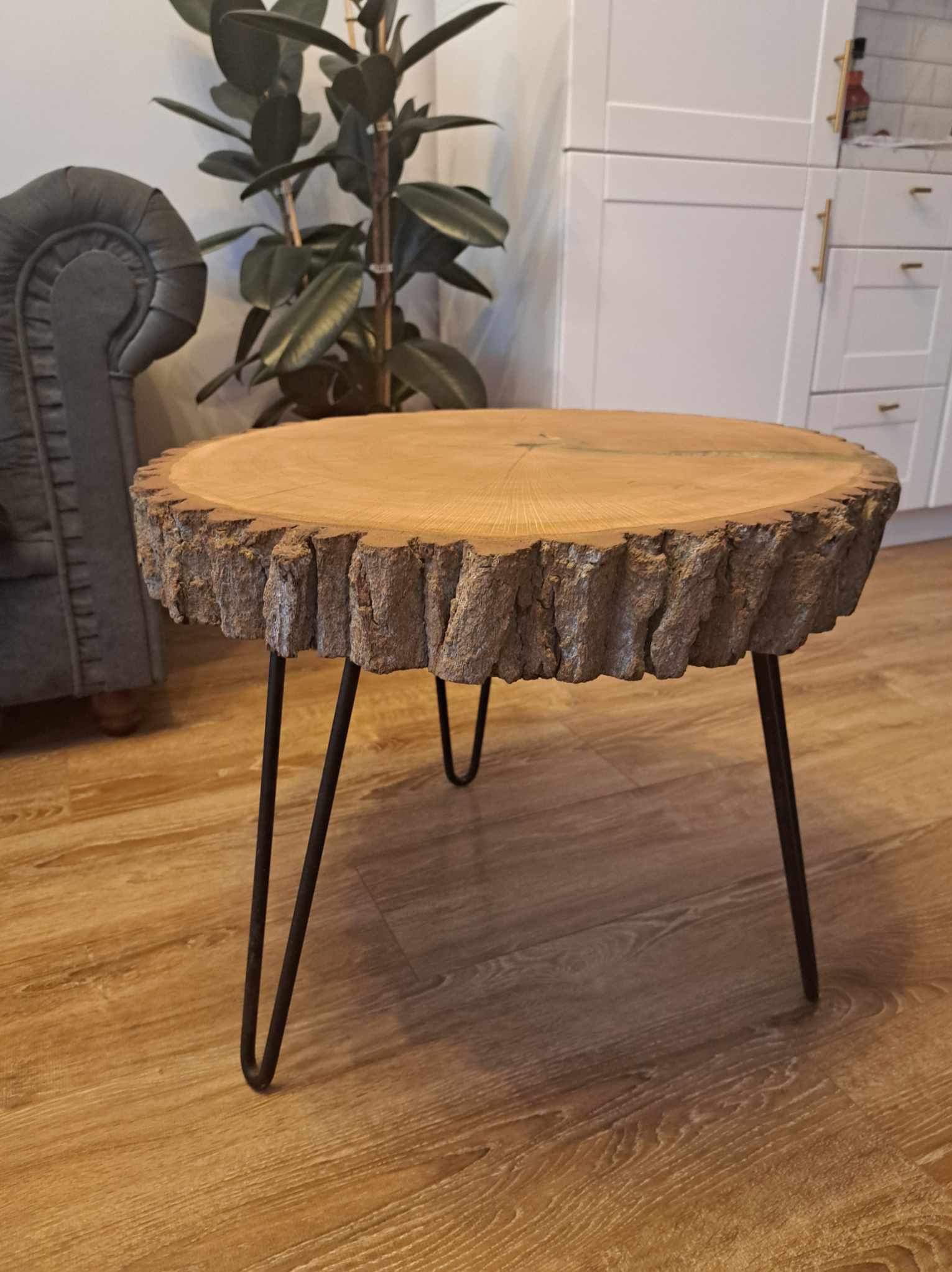 Stolik kawowy z blatem z plastra drewna z korą i metalowymi nogami w kształcie litery V, na drewnianej podłodze, w tle szary fotel i roślina doniczkowa.