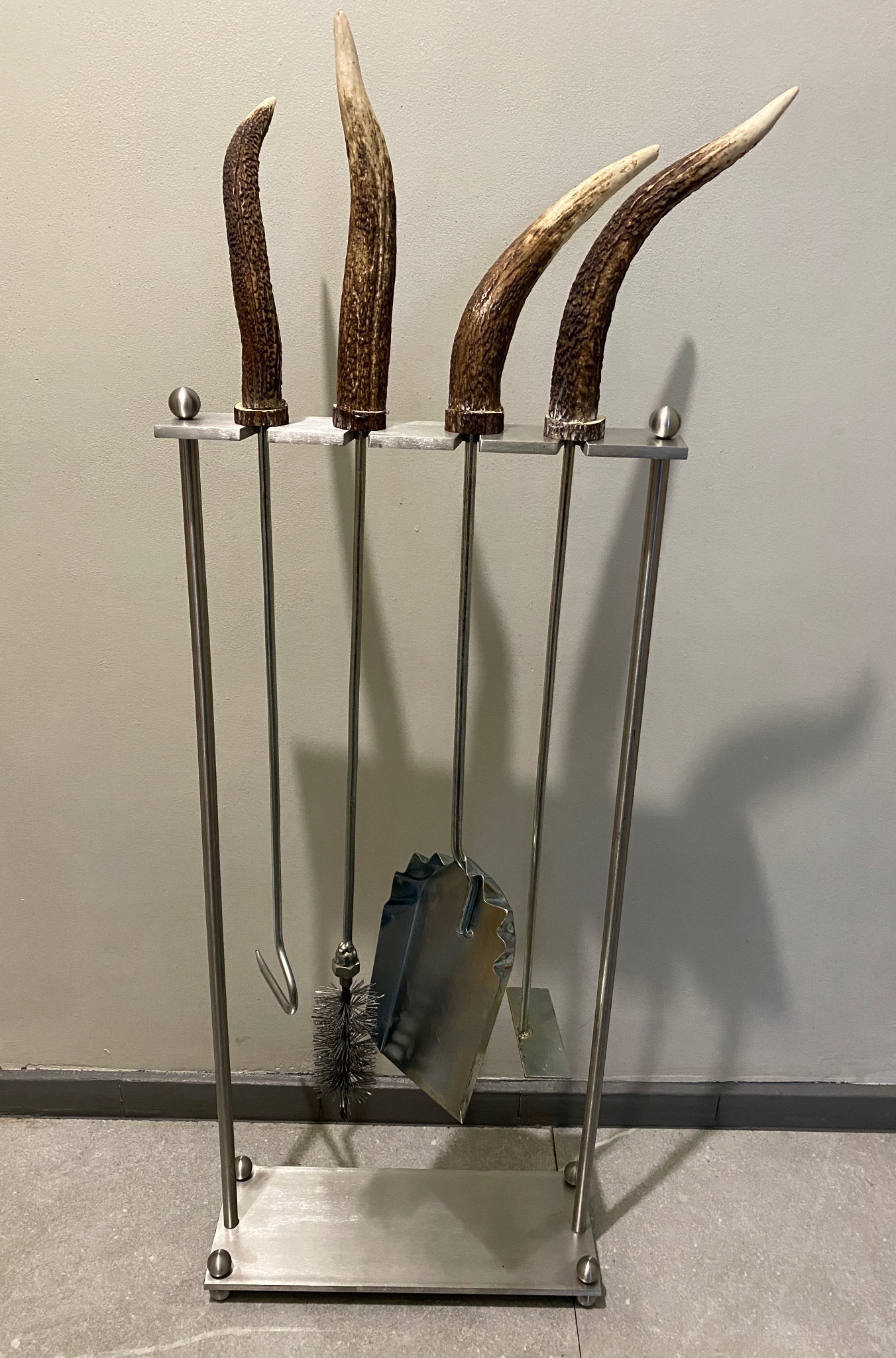 Zestaw kominkowy: pogrzebacz, szczotka i szufelka z uchwytami wykonanymi z poroża jelenia, umieszczone w metalowym stojaku o minimalistycznym designie.