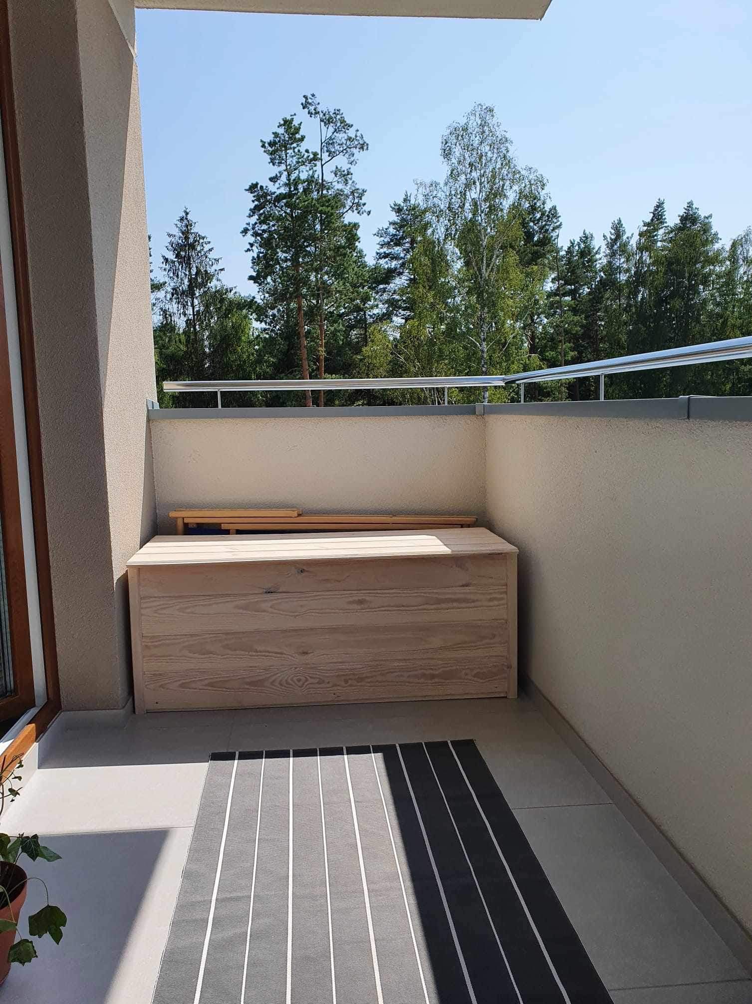 Drewniana skrzynia na balkonie z widokiem na las, z elementami do samodzielnego montażu na wierzchu, na szarym dywanie w paski.