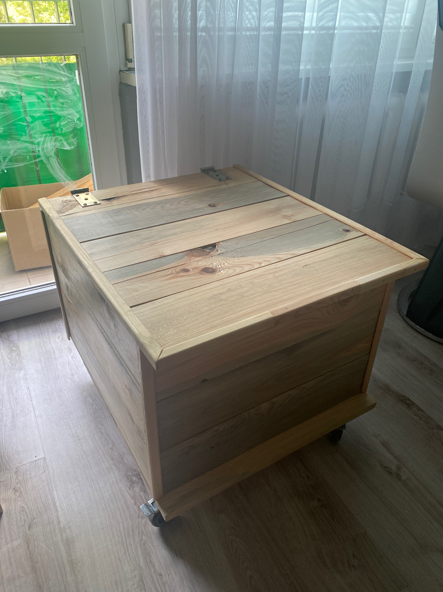 Drewniana skrzynia z wiekiem wykonanym z desek, zamontowana na kółkach, stoi na podłodze z paneli, widoczne zawiasy wieka i okno w tle.