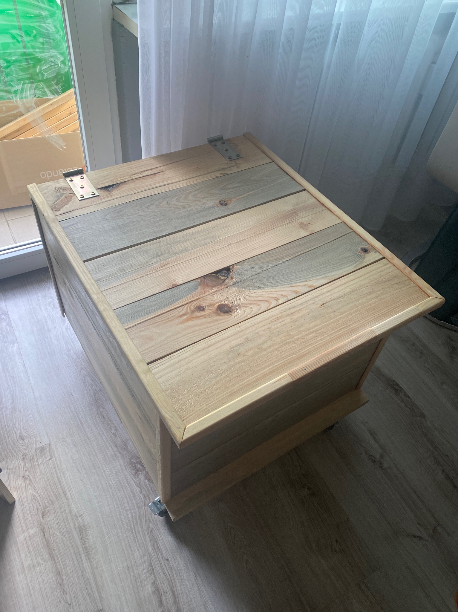 Drewniana skrzynia z wiekiem na zawiasach, wykonana z jasnych desek o zróżnicowanym odcieniu, umieszczona na kółkach, stoi na podłodze z paneli.