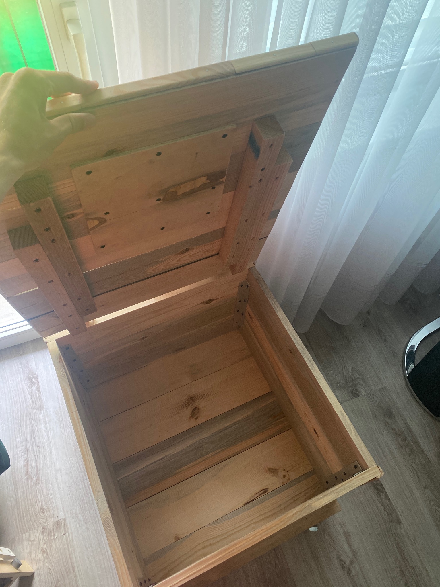 Otwarta, surowa, drewniana skrzynia z widocznymi elementami mocującymi, stojąca przy oknie z białymi zasłonami.