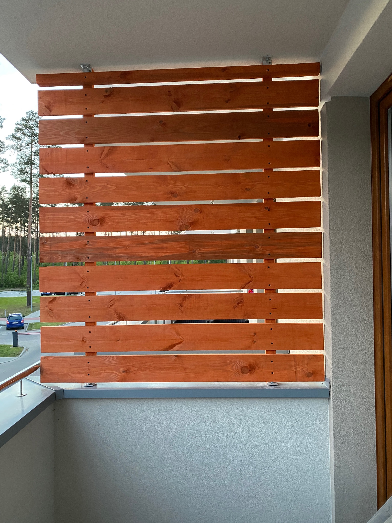 Drewniana przesłona balkonowa z poziomych desek w ciepłym odcieniu brązu, zamocowana do ściany i balustrady balkonu za pomocą metalowych uchwytów, z widocznymi przerwami między deskami...