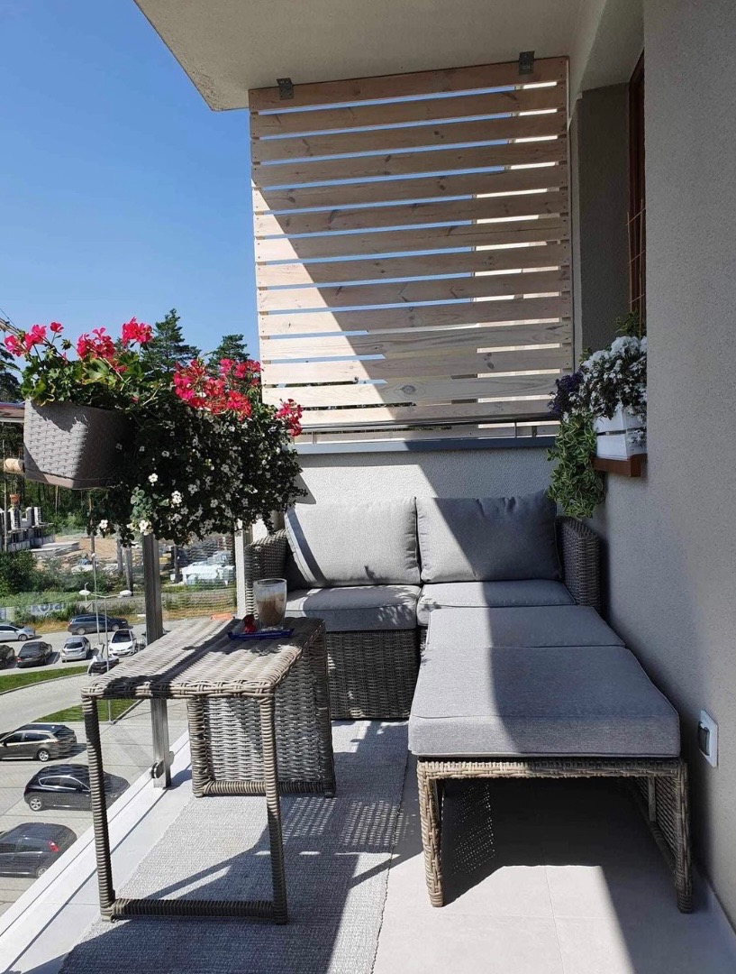 Zacieniony balkon z rattanowym narożnikiem, stolikiem i drewnianą osłoną, kwiaty w donicach, widok na ulicę z samochodami.