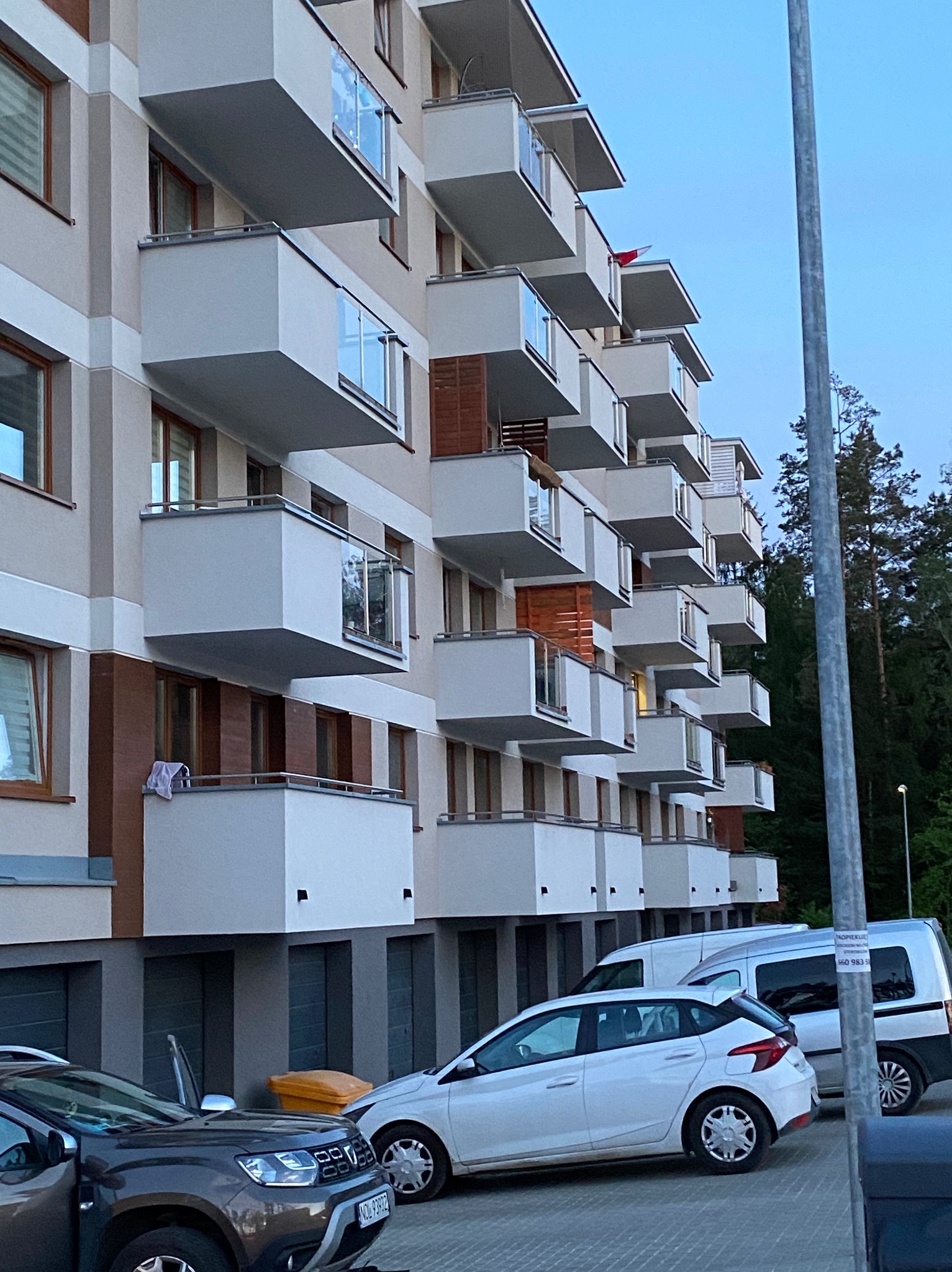 Nowoczesny budynek wielorodzinny z białymi, wystającymi balkonami z przeszklonymi balustradami, widok z dołu na fasadę z zaparkowanymi samochodami.