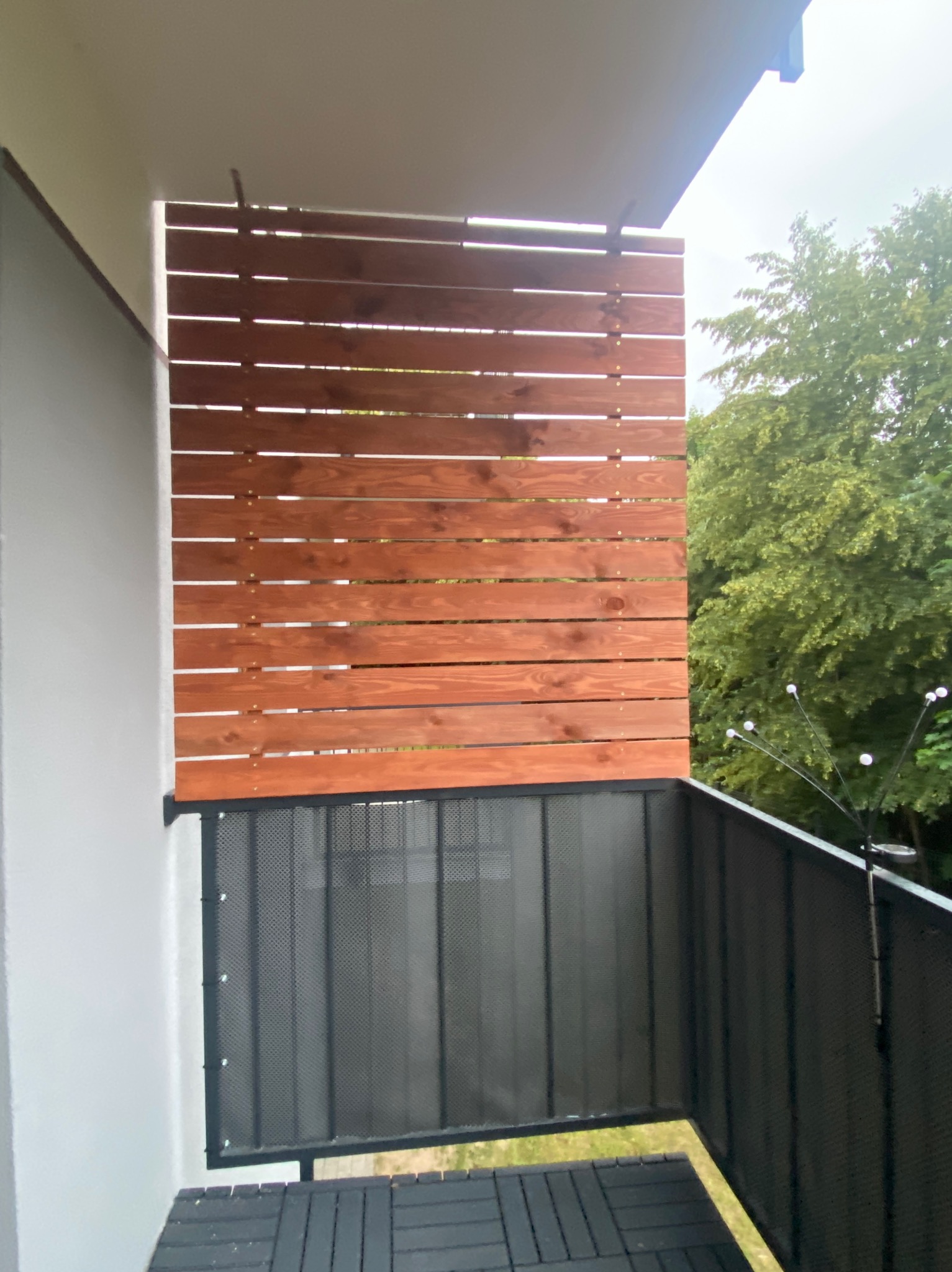 Drewniana osłona balkonowa z poziomych desek w ciepłym odcieniu brązu, zamontowana nad metalową balustradą z siatki, widok z balkonu na zieleń drzew.