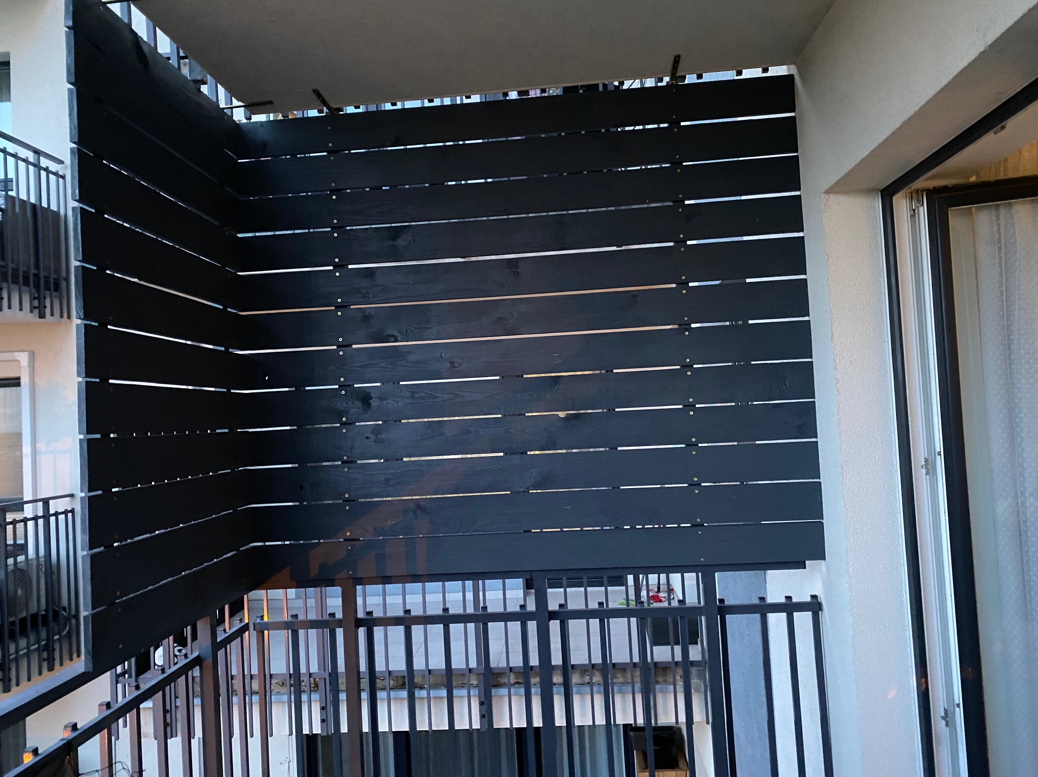 Częściowy widok balkonu z poziomymi, ciemnymi deskami tworzącymi przesłonę, zamontowanymi na metalowej konstrukcji balustrady.